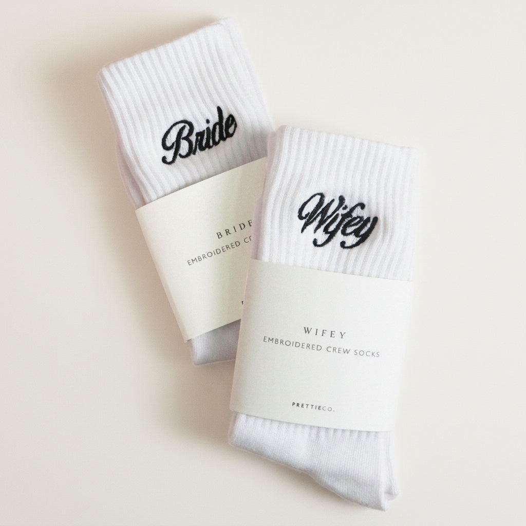 Two pairs of white socks with 'Bride' and 'Wifey' text on a pink background