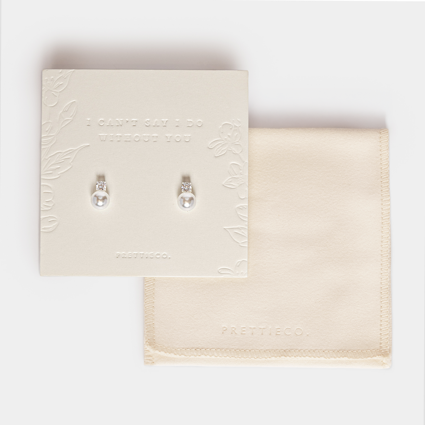 A pair of silver pearl earrings displayed on a cream-colored card, with a matching cream fabric pouch underneath on a light gray background. The card features embossed floral details and the text 'I CAN'T SAY I DO WITHOUT YOU'. Perfect for bridesmaid and bridal party gifting