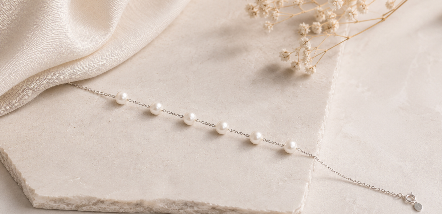 A delicate silver chain bracelet with small white pearl-like beads, draped elegantly across a light beige stone slab with soft cream fabric and dried flowers in the background.