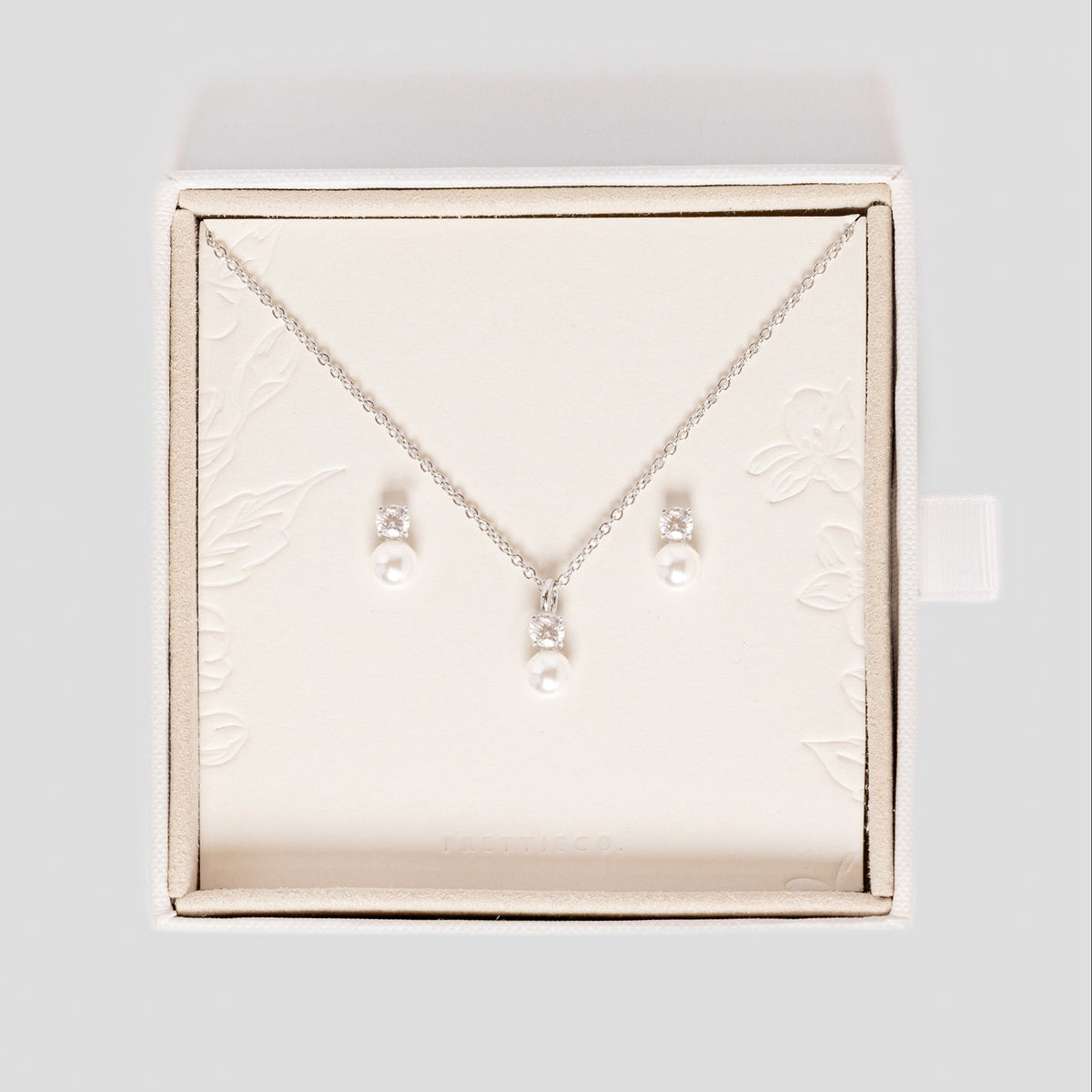 A silver necklace and matching stud earrings with small pearl-like beads, displayed inside an open cream-colored jewelry box on a light gray background. Perfect for wedding day bridal jewellery 