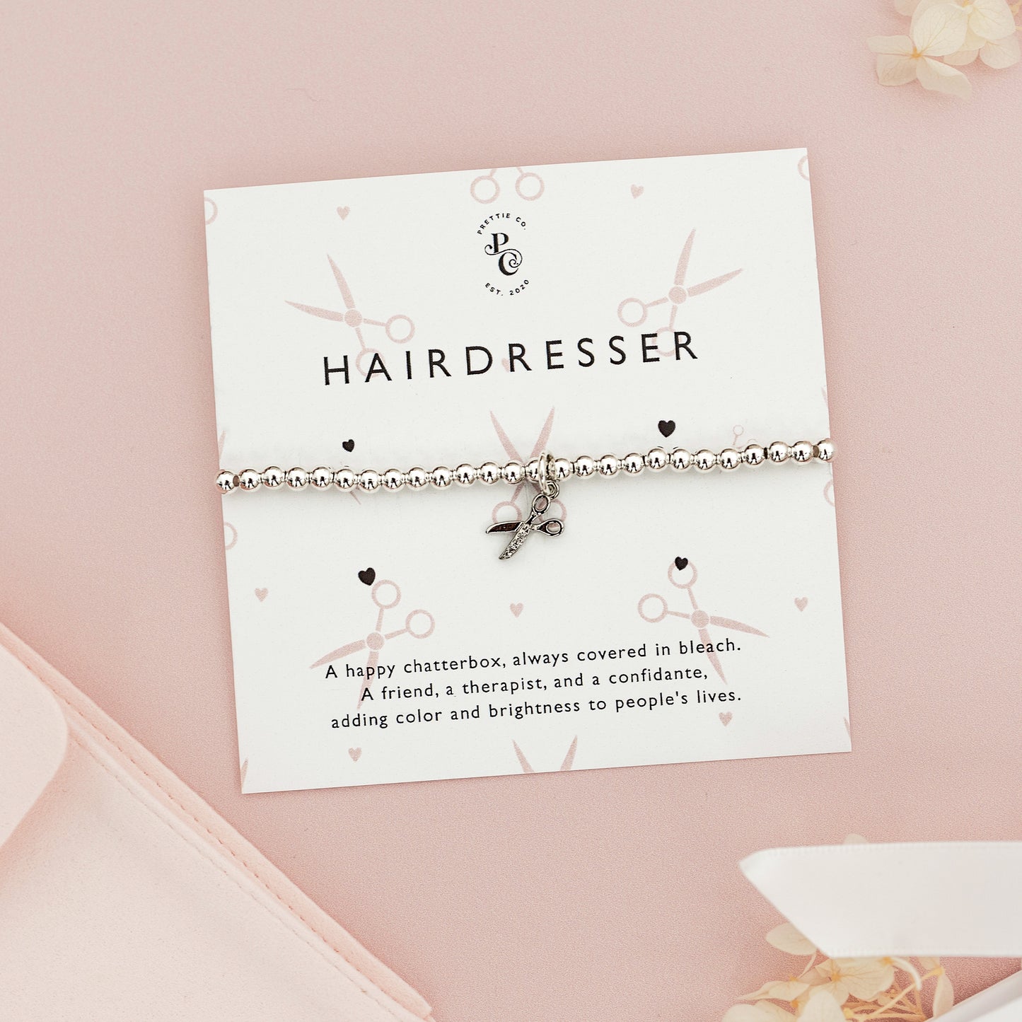 Personalised silver beaded bracelet with a scissors charm for hairdressers, displayed on a custom card with playful scissor and heart illustrations, set against a pink background.