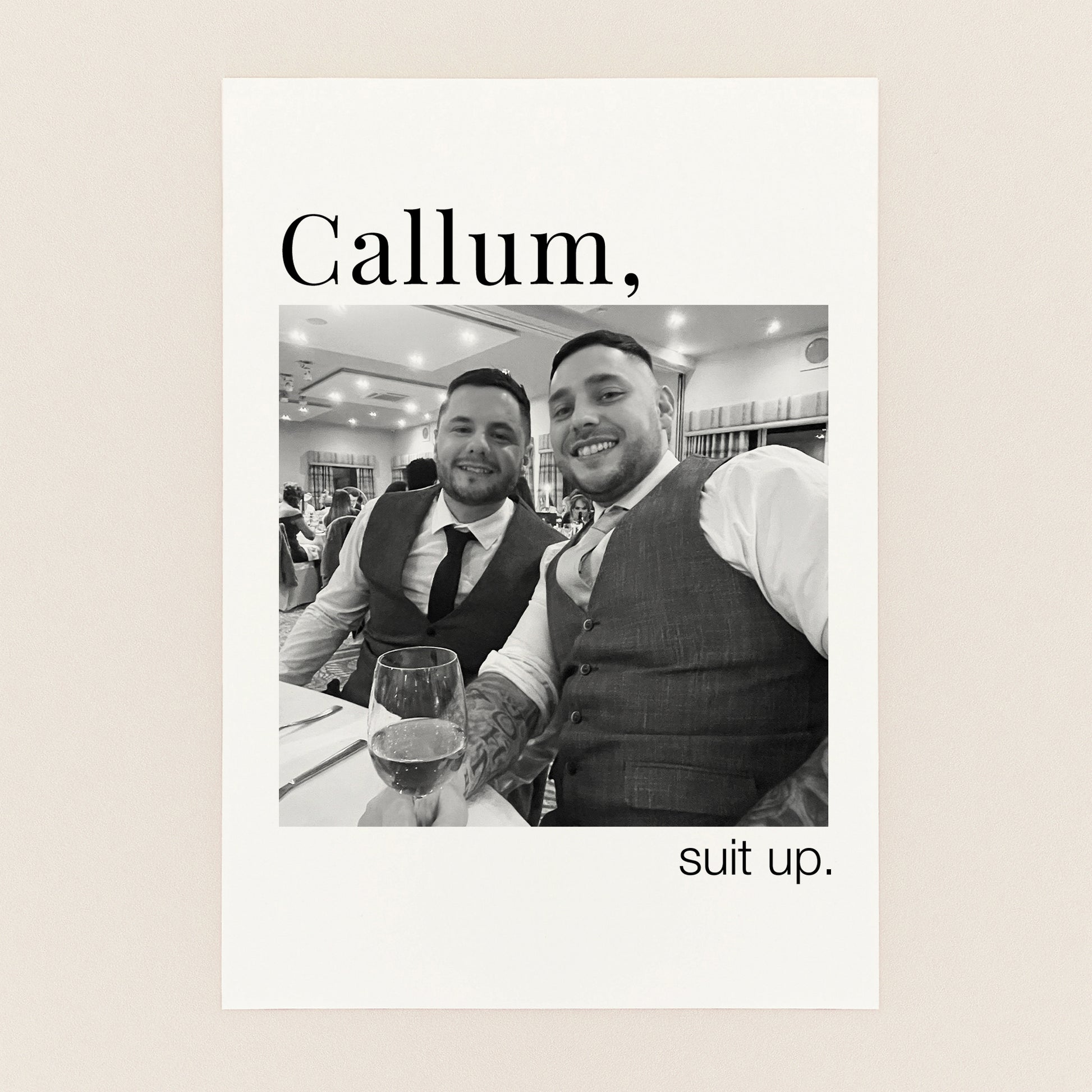 A personalized black and white wedding print featuring a photo of two men in suits, with the text 'Callum,' at the top and 'suit up.' at the bottom, displayed on a light beige background.