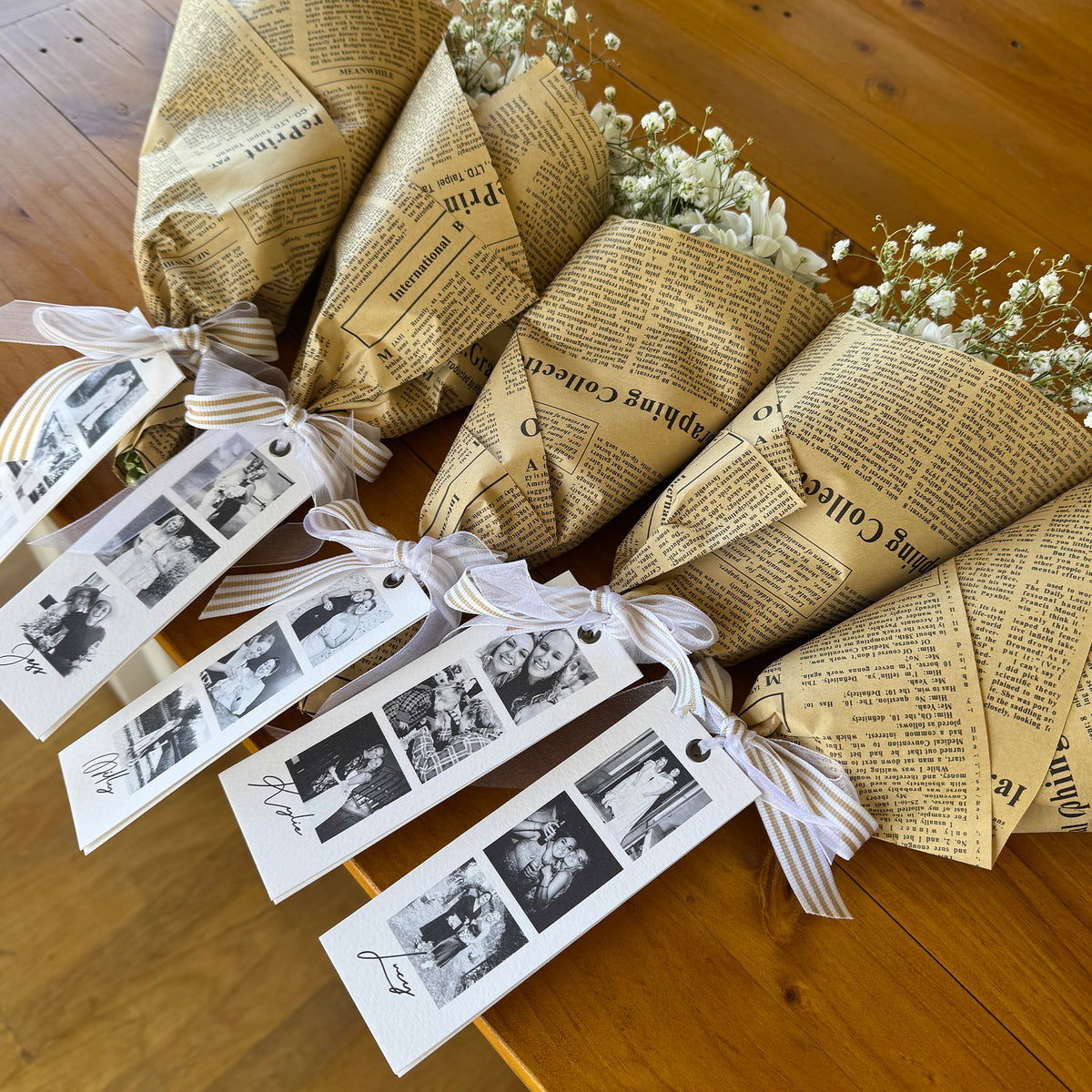Five wrapped flower bouquets tied with striped ribbons and attached white photo tags, arranged on a wooden tabletop. The bouquets are wrapped in newspaper-style paper with printed text, and small white baby's breath flowers are visible at the top.