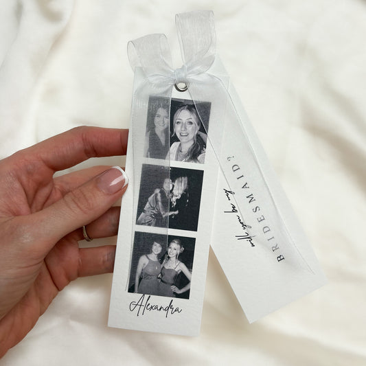 A hand holds a white bridal shower favor tag tied with a sheer white ribbon. The tag features three black-and-white photos of a bride and groom, the name 'Alexandra' in script, and a second attached tag reading 'BRIDESMAID' with a signature.