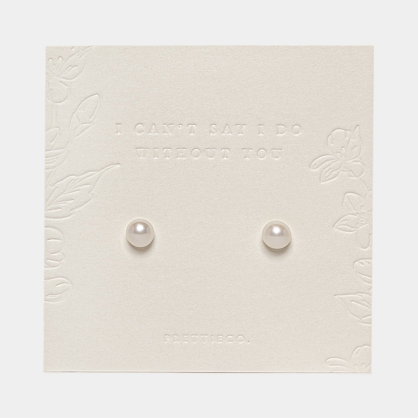 A pair of small round pearl stud earrings displayed on an off white-colored embossed card with floral borders and the text 'I CAN'T SAY I DO WITHOUT YOU' and 'FRETTIECO.'.