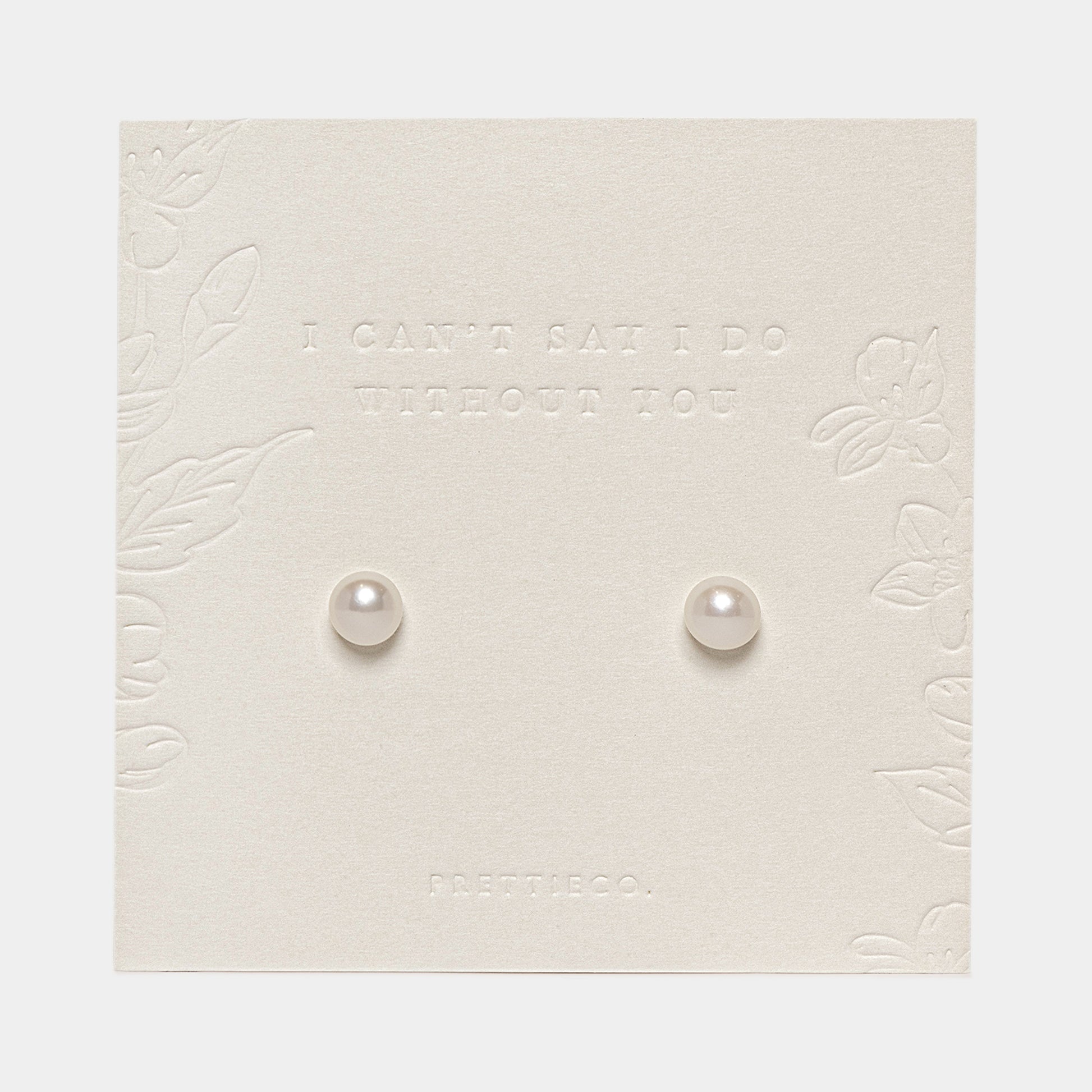 A pair of small round pearl stud earrings displayed on an off white-colored embossed card with floral borders and the text 'I CAN'T SAY I DO WITHOUT YOU' and 'FRETTIECO.'.